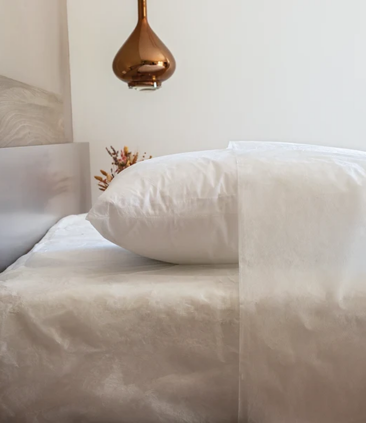 Lit habillé de linge jetable blanc, oreiller et drap en non-tissé, avec une suspension cuivrée et un bouquet séché en arrière-plan