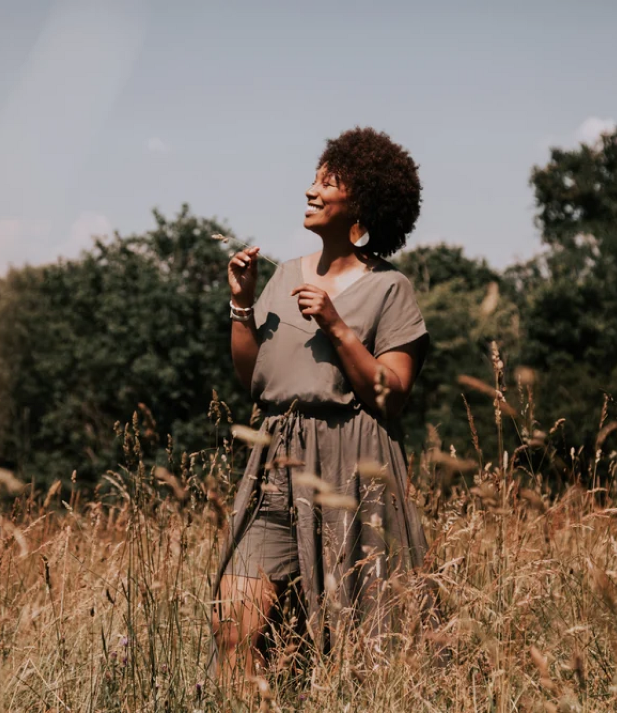 Femme souriante aux cheveux bouclés, en robe kaki, tenant un brin d'herbe dans un champ de hautes herbes dorées