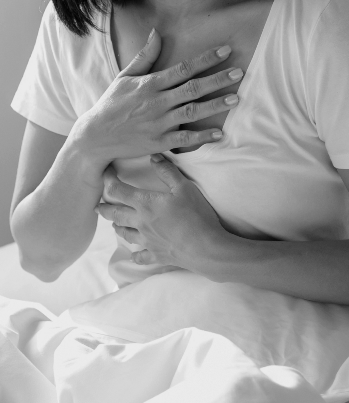 Femme en t-shirt blanc assise dans un lit, les mains posées sur la poitrine, photographie en noir et blanc