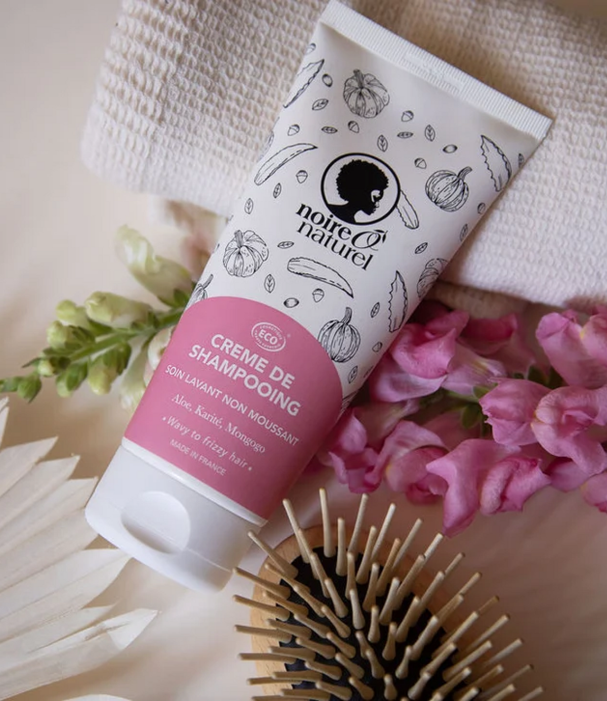 Tube de crème de shampooing Noire O Naturel posé près de fleurs roses, d'une serviette gaufrée et d'une brosse en bois